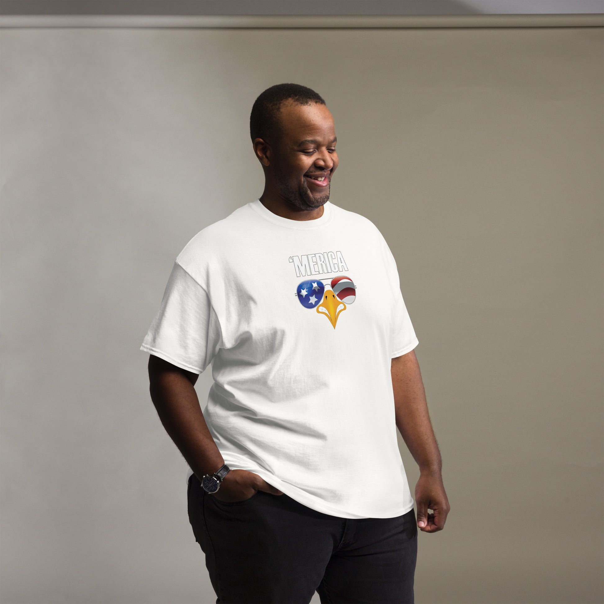 The image portrays a middle-aged African American man smiling and standing casually. He is wearing a white t-shirt that features a playful graphic design with the text "MERICA" and an eagle's beak wearing sunglasses that reflect the American flag—stars on a blue background on one lens and red and white stripes on the other. The man's relaxed demeanor, combined with the humorous patriotic shirt, creates a light-hearted and approachable image.