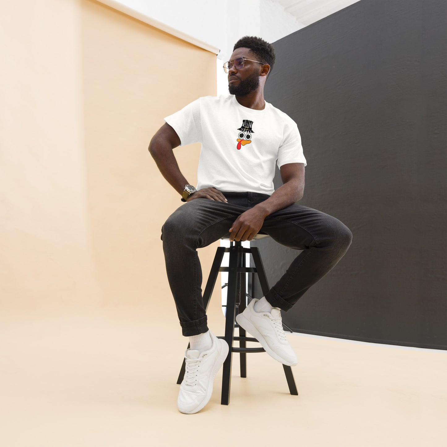 A man with a neatly trimmed beard and glasses is wearing a white t-shirt featuring a playful turkey graphic. The turkey has cartoonish eyes, an orange beak, and a red snood, along with a black hat that reads "TURKEY MASTER" in bold letters. He is sitting on a stool with one hand resting on his thigh and the other on the stool's leg, creating a relaxed and confident pose. He is wearing black jeans and white sneakers, and the background is a minimal studio setting with beige and black walls.