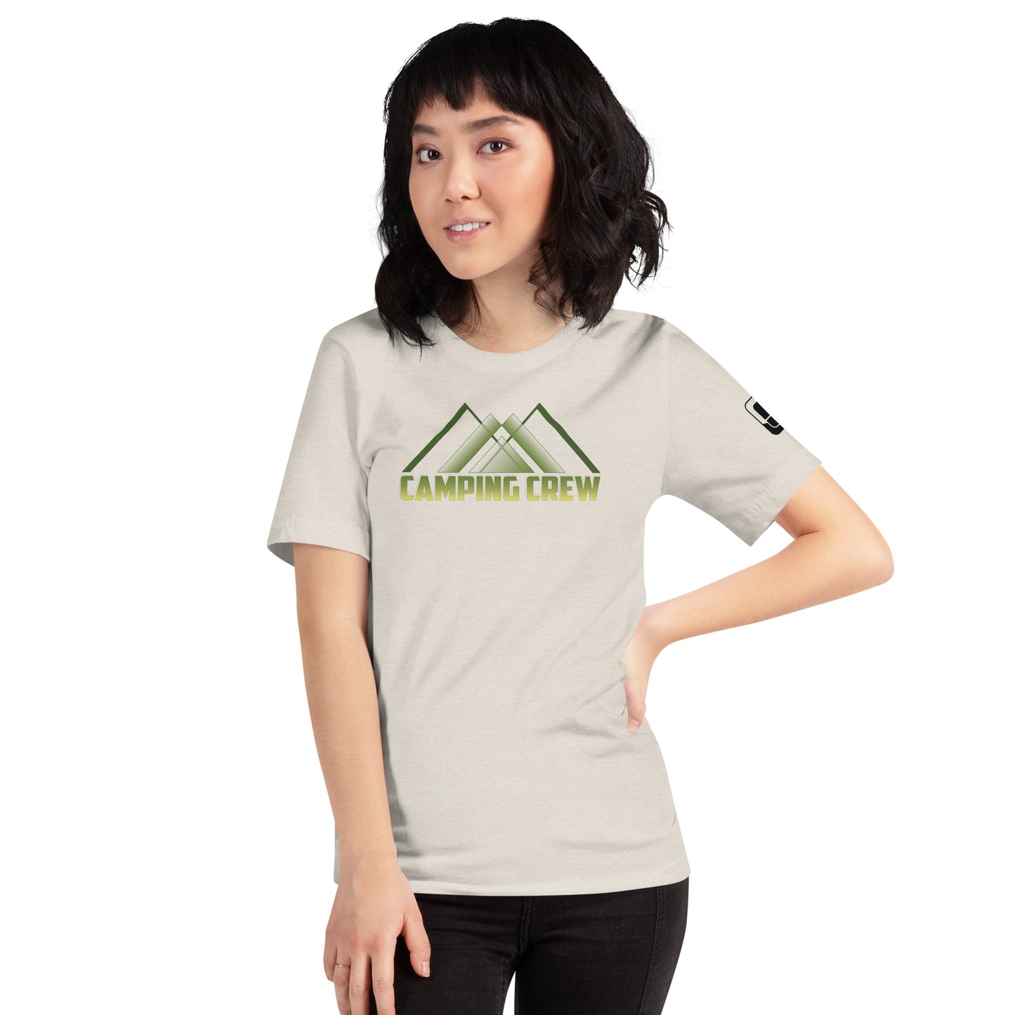 An East Asian woman with short, wavy black hair models a light grey t-shirt that prominently features the green 'CAMPING CREW' logo with a stylized mountain design. She poses with one hand on her hip, giving a subtle, playful look towards the camera. The shirt's casual style is accented with a small logo patch on the sleeve, highlighting her adventurous spirit.