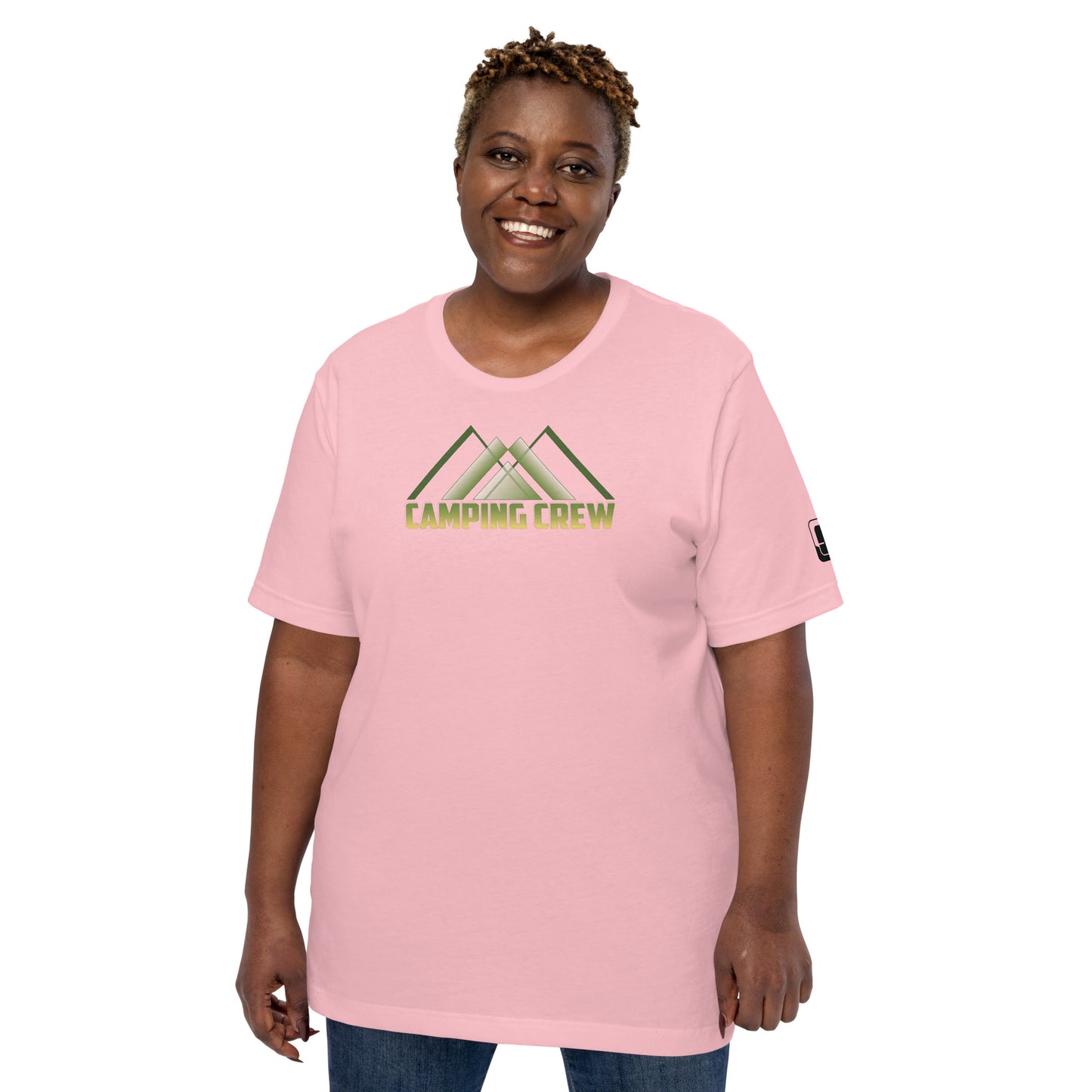 An African American woman with short, curled hair and a warm smile models a soft pink t-shirt. The shirt features a prominent green 'CAMPING CREW' logo with an abstract mountain design above the text. Her pose is friendly and relaxed, emphasizing the casual, inviting look of the t-shirt suitable for outdoor activities.