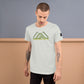 A young Caucasian man with short brown hair, and tattoos on both arms models a light grey t-shirt that features green to yellow text reading  ‘Camping Crew’  below an abstract mountain design. He stands with his head slightly bowed, looking down contemplatively against a dual-tone background of blue and warm beige, giving the photo a serene, introspective mood.