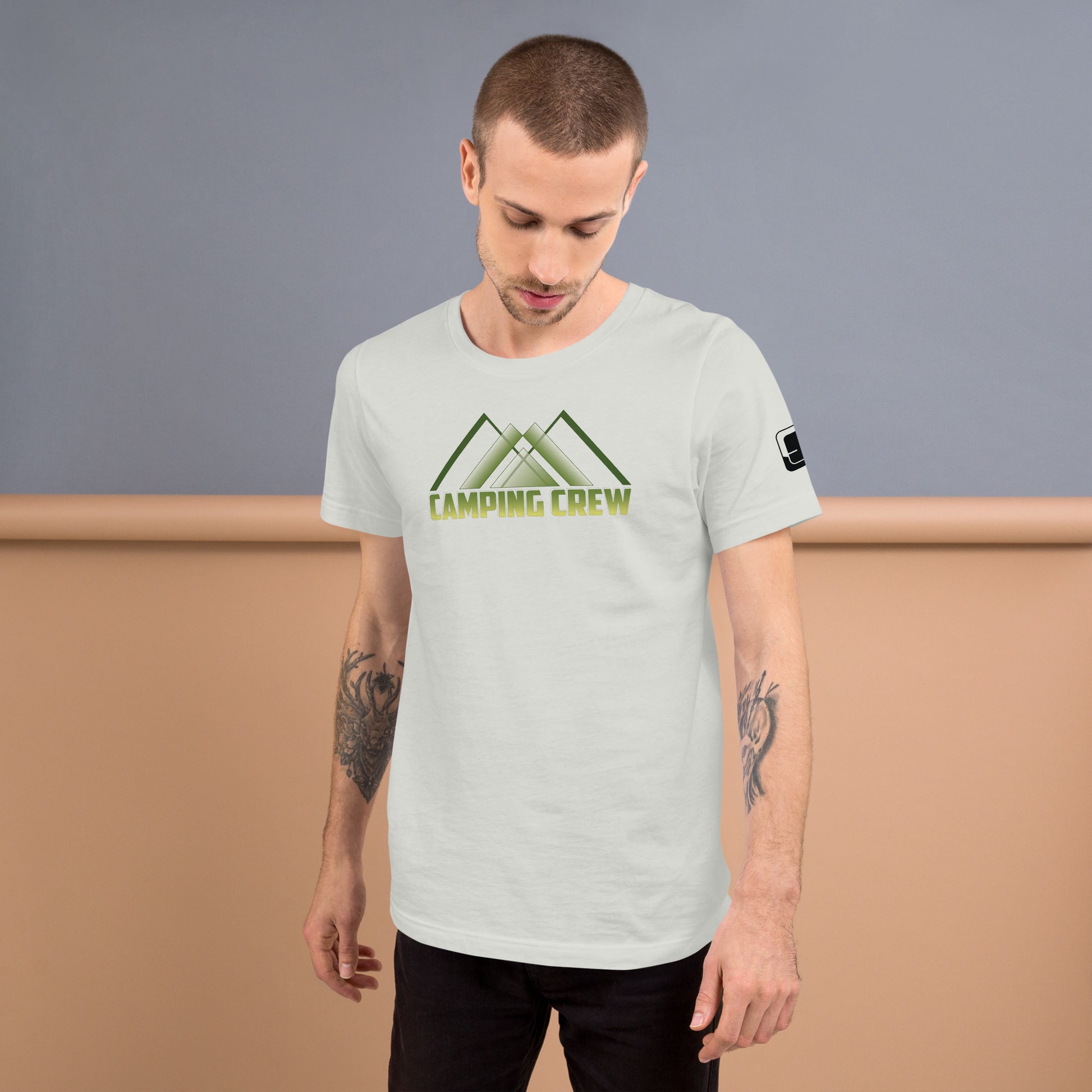 A young Caucasian man with short brown hair, and tattoos on both arms models a light grey t-shirt that features green to yellow text reading  ‘Camping Crew’  below an abstract mountain design. He stands with his head slightly bowed, looking down contemplatively against a dual-tone background of blue and warm beige, giving the photo a serene, introspective mood.