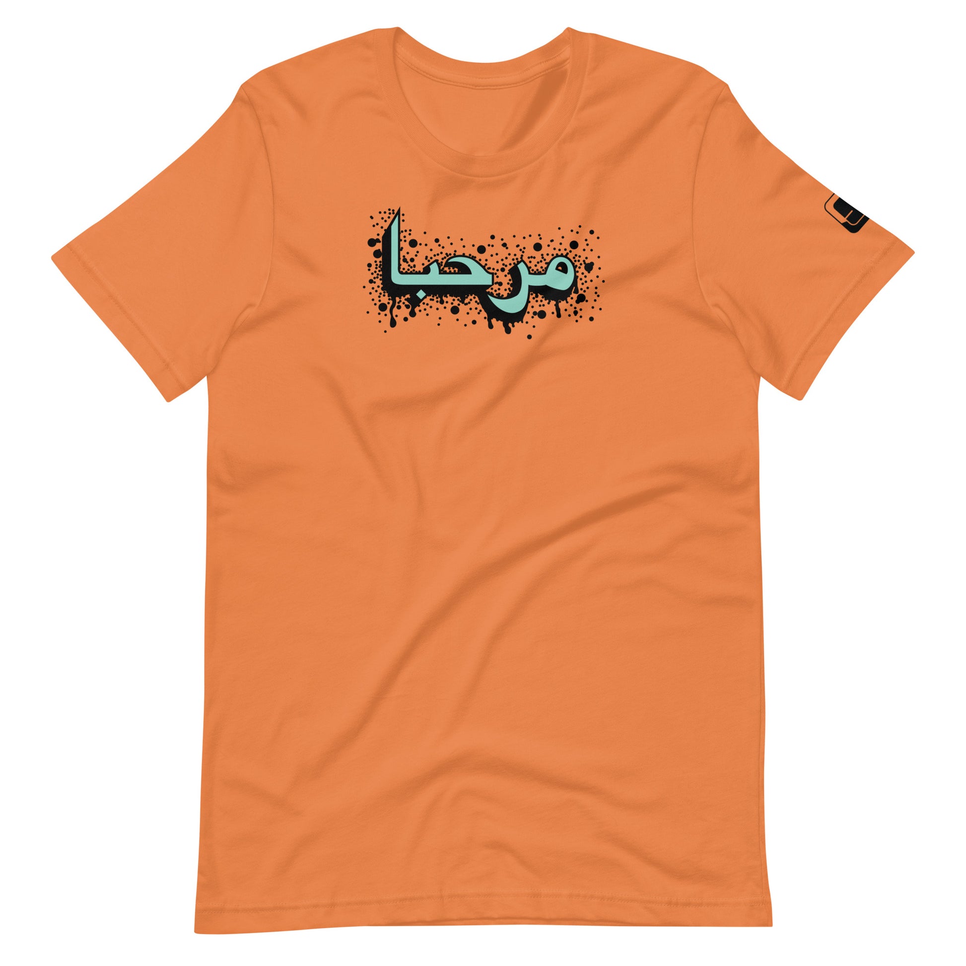 Burnt orange t-shirt featuring central turquoise Arabic calligraphy with black shadow and scattered ink dots, accompanied by a small black logo patch on the sleeve, displayed against a white background.