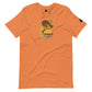 An orange-colored t-shirt laid flat, adorned with a "Taco Tuesday" graphic. The design showcases a playful fish in a taco, complete with a colorful sombrero, happily giving a thumbs up. The graphic's background is a vibrant yellow, with the words "Taco Tuesday" in bold, colorful letters, evoking a festive and lively spirit. The overall look is casual and fun, perfect for a themed event or casual wear.