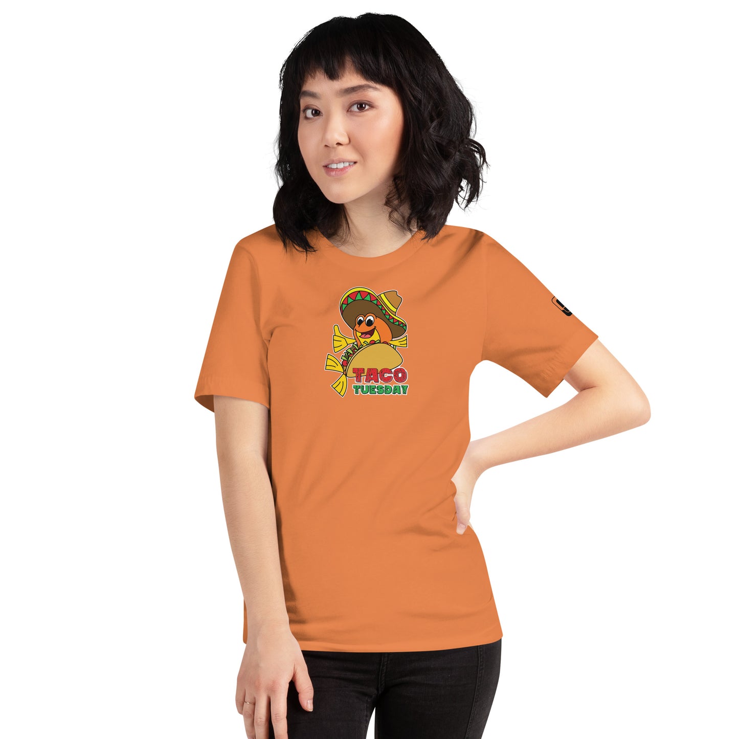 A young Asian woman with shoulder-length black hair, standing slightly turned to the side, wearing a orange-colored t-shirt. The t-shirt displays a playful "Taco Tuesday" graphic featuring a fish in a taco, adorned with a sombrero, and giving a thumbs up. Her casual stance and the soft smile suggest a relaxed and approachable vibe, complementing the fun and whimsical theme of her shirt.