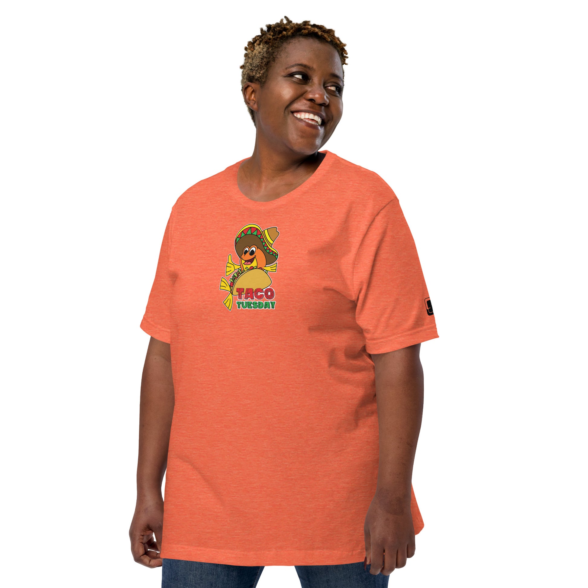 A joyful African American woman with a bright smile, wearing an orange colored t-shirt featuring a playful "Taco Tuesday" design. The shirt's graphic shows a cheerful fish in a taco, wearing a sombrero and giving a thumbs-up. Her relaxed and happy demeanor, paired with casual denim jeans, complements the light-hearted, festive theme of the shirt, making it a charming choice for casual wear or themed events.