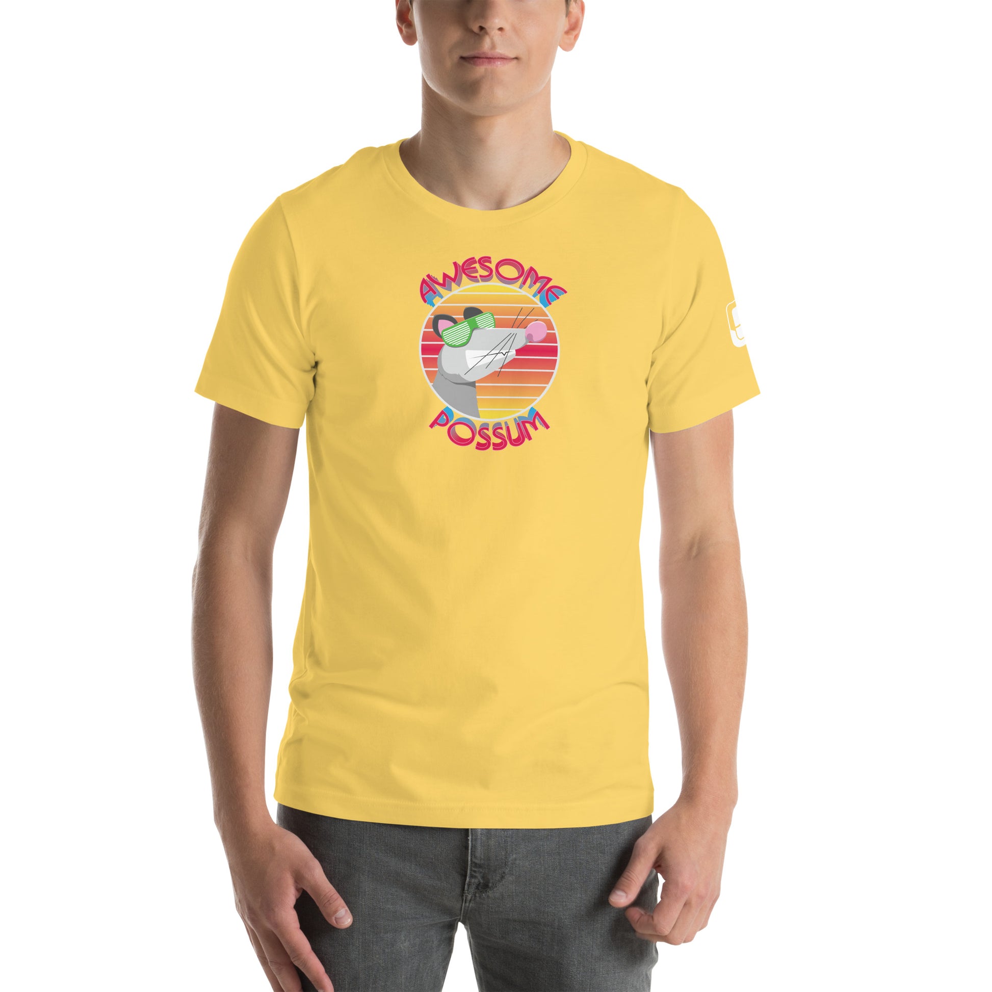 A young man is shown wearing a vibrant yellow t-shirt adorned with a fun graphic design. The design features a cartoon possum's head in sunglasses, encircled by the phrases "AWESOME" and "POSSUM" in colorful, bold text against a backdrop of pink and orange sunset stripes. The playful imagery is centered on the shirt, conveying a casual and lighthearted aesthetic.