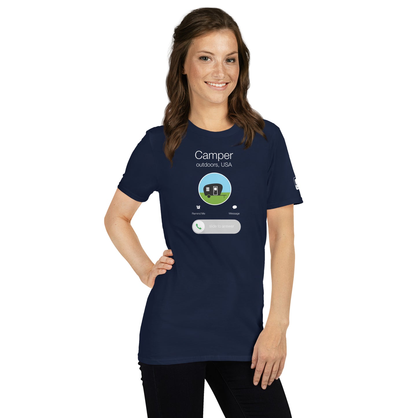 A Caucasian woman with medium-length brown hair and a friendly smile models a navy blue t-shirt featuring a unique smartphone-inspired graphic. The t-shirt displays a dark blue circle centered on the chest with the design of a simple black camper trailer against a blue and green background. Above the graphic, the text reads "Camper outdoors, USA." Below the circle, options include "Remind Me," "Message," and a slider labeled "slide to answer," creatively blending outdoor camping themes with a modern, techy 