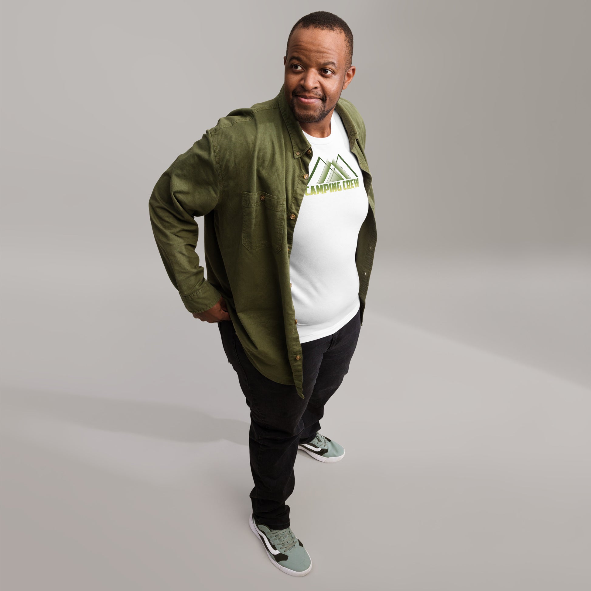 A confident African American man models a casual ensemble featuring a white t-shirt under a green buttoned shirt jacket. The t-shirt is emblazoned with a green and white 'Camping Crew' design depicting an abstract mountain. He stands with one hand on his hip and the other slightly adjusting his jacket, smiling broadly, wearing dark pants and light green sneakers, against a soft grey background