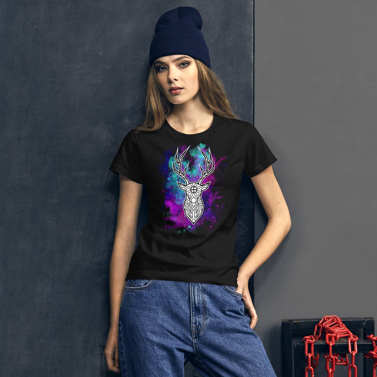 A young Caucasian woman wearing a dark blue beanie models a black T-shirt featuring a vivid geometric deer head design. The deer is accentuated by a colorful nebula background in shades of purple, blue, and pink. She stands with one hand on her hip, leaning slightly against a dark blue wall in a casual yet stylish pose, with an intense gaze towards the camera.