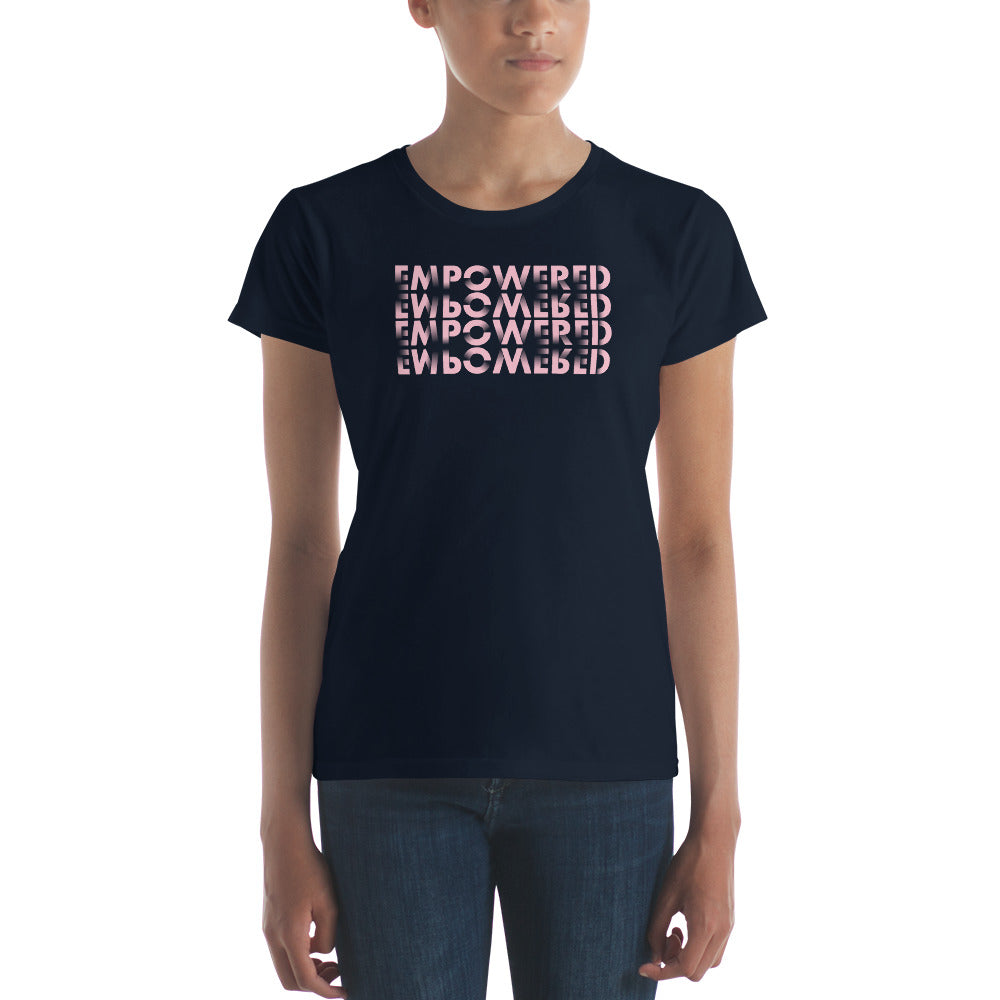 
A young woman stands with a neutral expression, wearing a dark navy t-shirt featuring the word "EMPOWERED" repeated four times in a layered design. The text transitions in color from dark to light pink, providing a striking contrast against the shirt's dark background. The t-shirt fits snugly, complementing her casual style paired with blue jeans. She maintains a relaxed posture, with her arms naturally by her sides, embodying a subtle confidence.