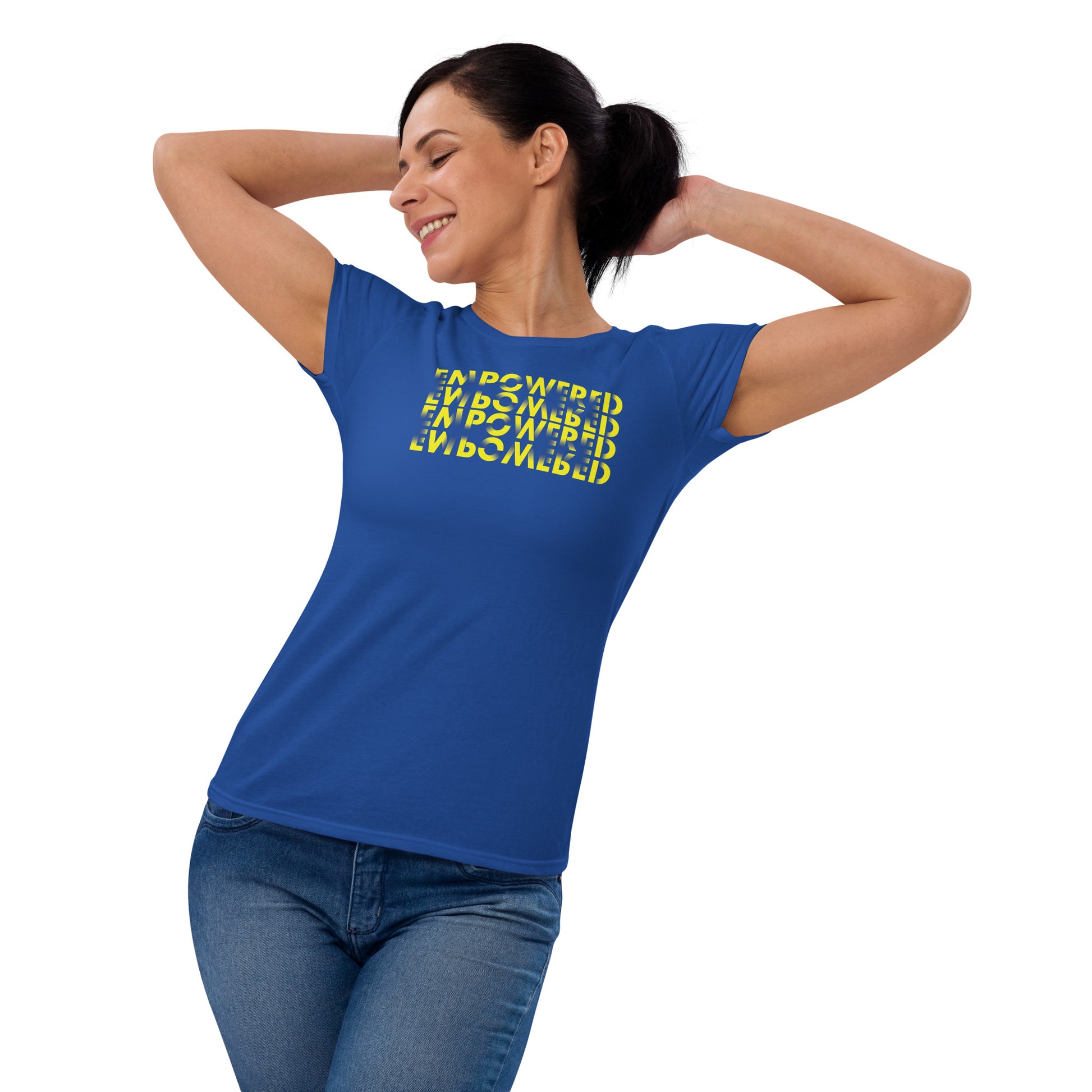 
A middle-aged Caucasian woman in a playful pose, wearing a royal blue t-shirt that features a complex text design. The shirt's design cleverly uses the word "EMPOWERED" printed four times in varying orientations to form a cube-like visual effect, with the text in yellow against the darker shirt. She stands with her hands confidently lifting her hair up, smiling gently, enhancing the empowered message of her attire.